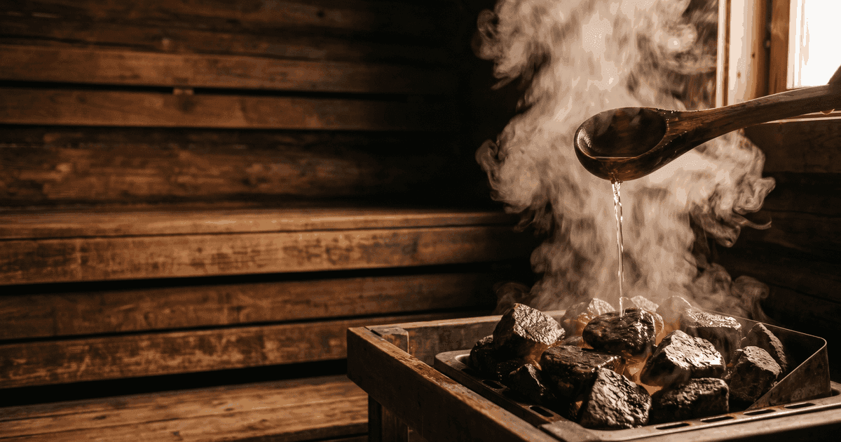 A wooden ladle pouring water onto glowing sauna stones, with a soft burst of steam rising into a warmly lit Finnish sauna.